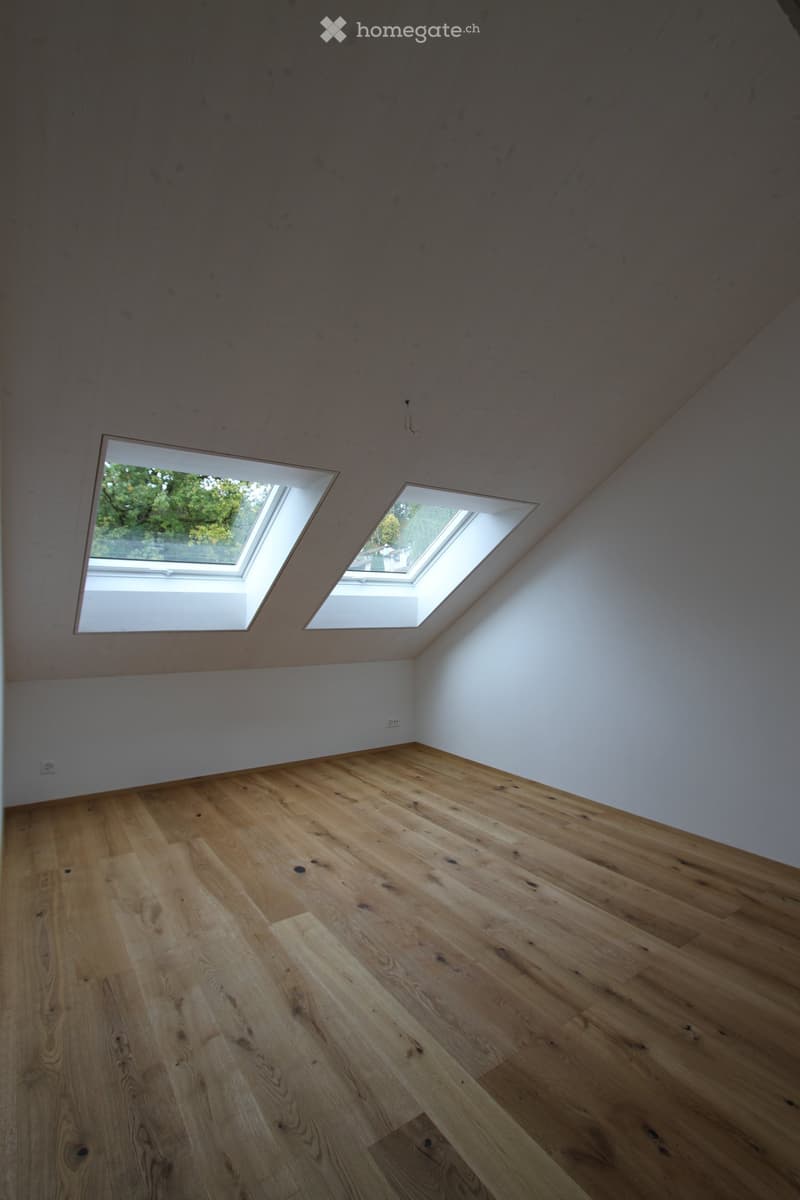 Empty attic room with wooden flooring and two skylight windows, white walls, and exposed wooden beams on the ceiling.