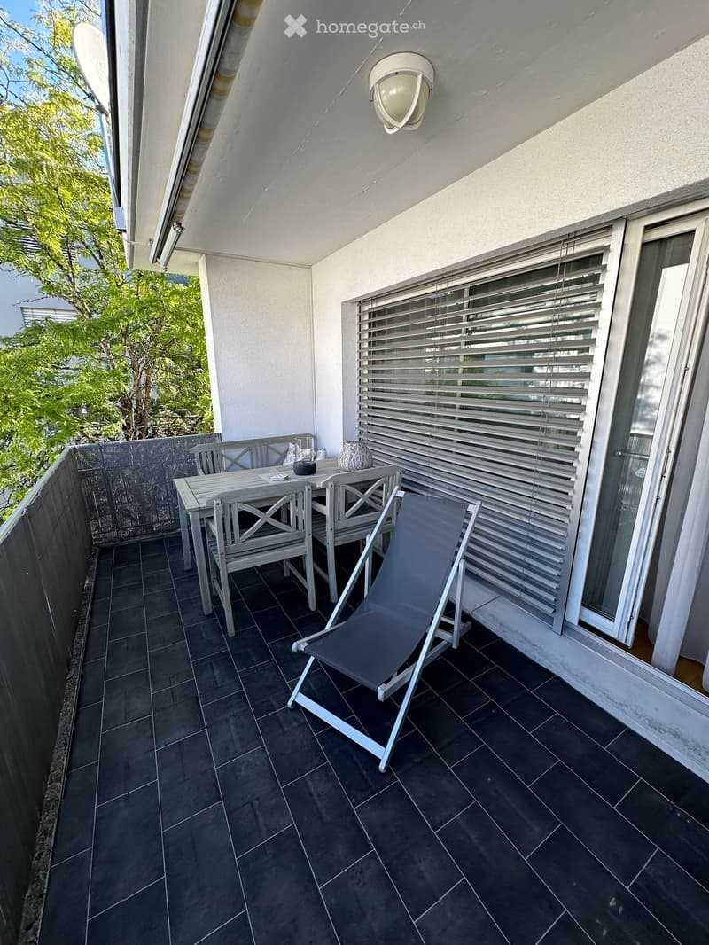 Balcony with dark tiled floor, white table, two chairs, and a lounge chair, with a view of greenery.