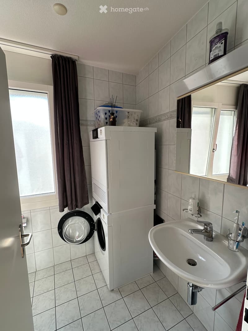 Bathroom with a white sink, faucet, toothbrush holder, washing machine, mirror, and a curtain on the window. The floor is tiled, and there is a basket on top of the washing machine.