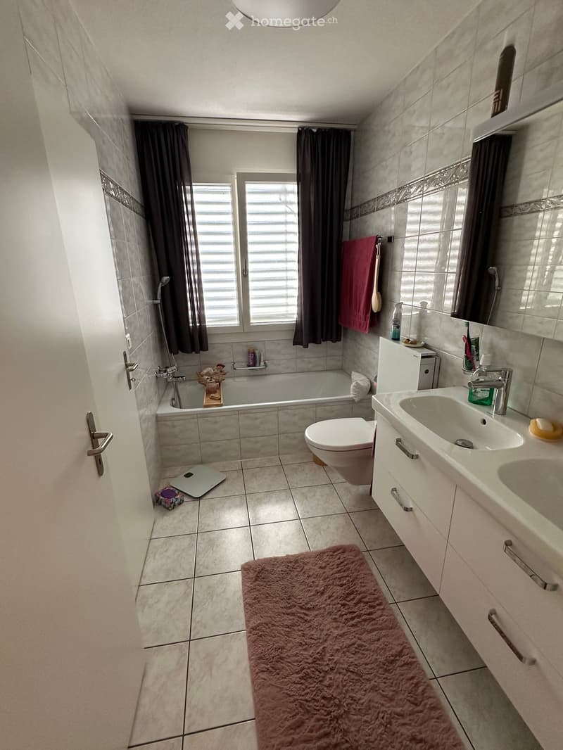 Modern bathroom with a bathtub, toilet, double sink, mirror, and a pink bath mat. The room features tiled walls and floors, a window with blinds, and a shower area with a curtain.