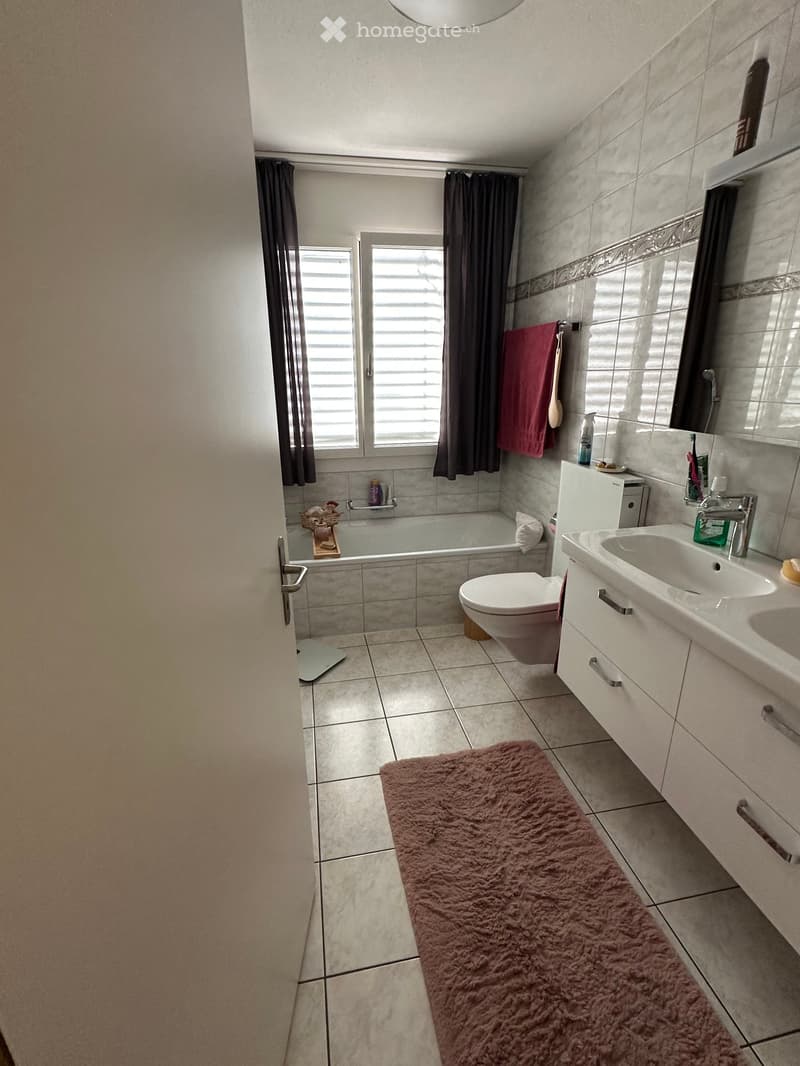 Modern bathroom with a bathtub, toilet, sink, mirror, and a pink towel on the wall. The room features tiled walls and floor, a window with dark curtains, and a fluffy pink rug on the floor.