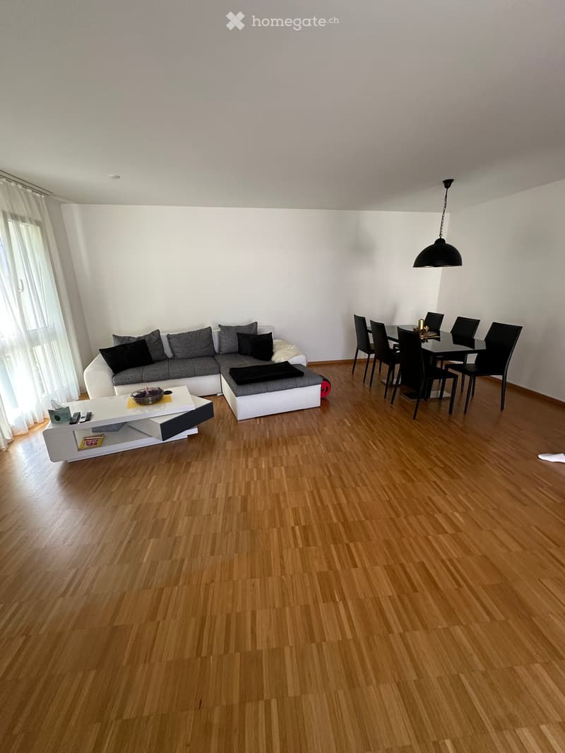 Modern living and dining area with light wooden flooring, white walls, and large windows with white curtains. Features a grey sectional sofa, white coffee table, and a black dining table with chairs.
