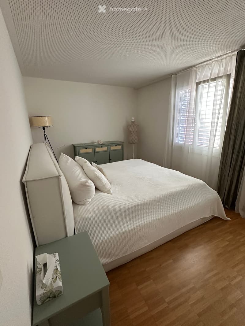 Modern bedroom with a large bed, nightstand, lamp, and a small drawer unit. The room features wooden flooring and large windows with blinds.