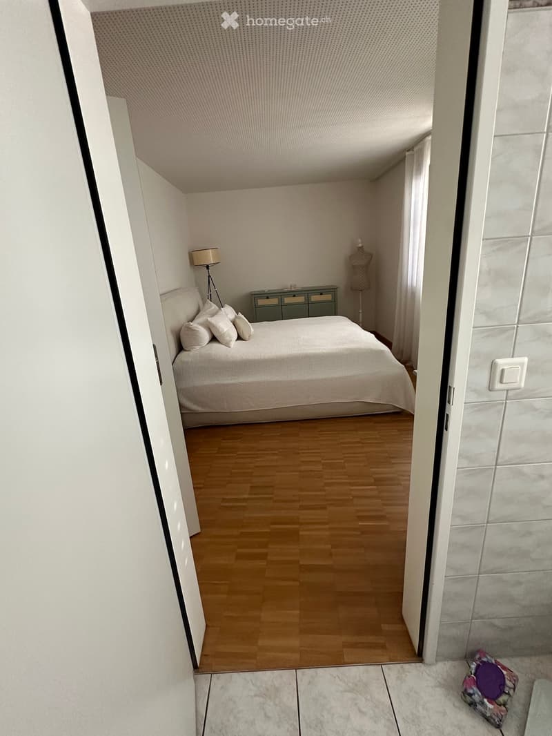 Modern bedroom with a large bed, white bedding, nightstand, lamp, and a small drawer unit. Wooden flooring and white walls.