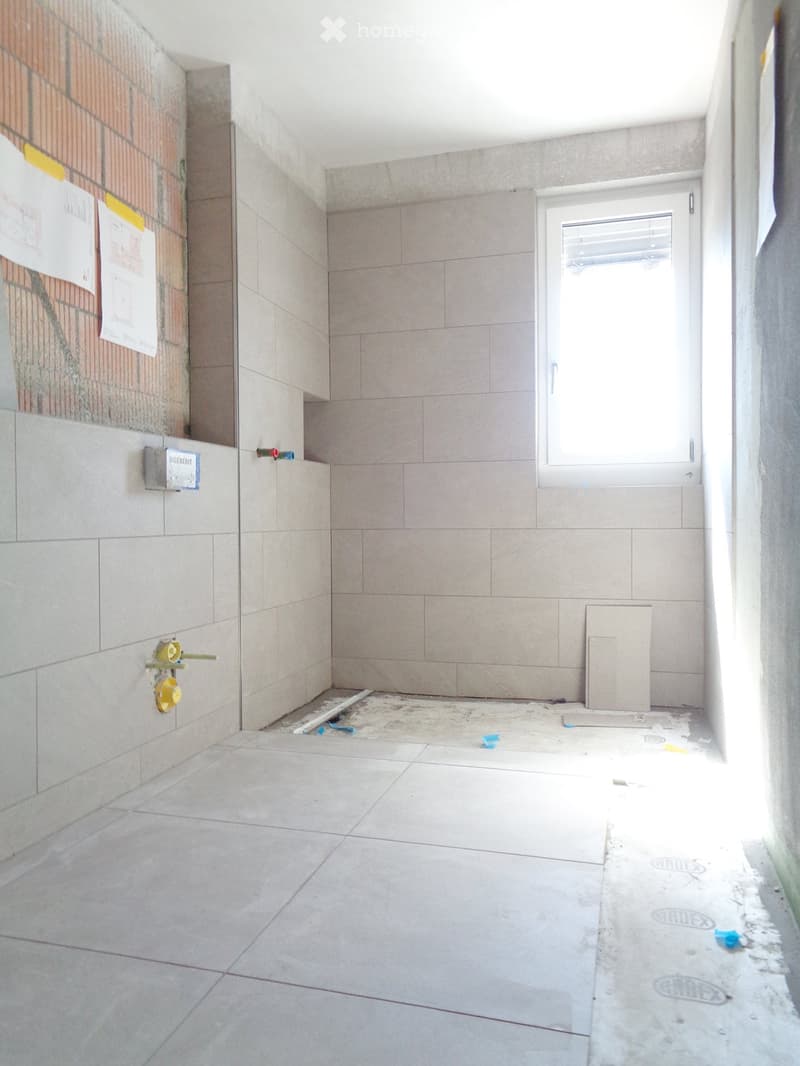 Empty room with tiled walls and floor, a window with blinds, and construction materials scattered on the floor.
