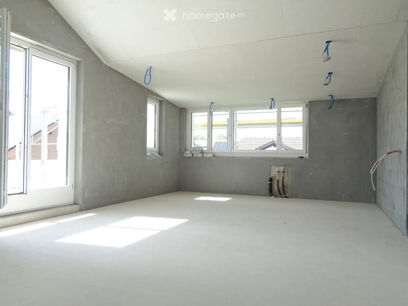 Empty room with grey walls, white ceiling, and concrete floor. Windows and door provide natural light. Exposed electrical outlets and pipes are visible.