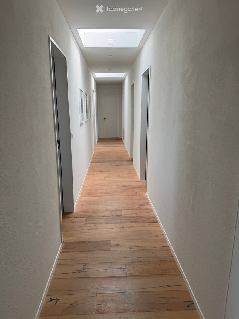 A long, narrow hallway with wooden flooring and white walls, featuring several doors and a skylight at the far end.