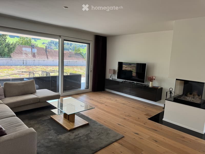 Modern living room with wooden flooring, large windows offering a scenic view, a grey sectional sofa, a glass coffee table, a flat-screen TV on a black cabinet, and a contemporary fireplace.