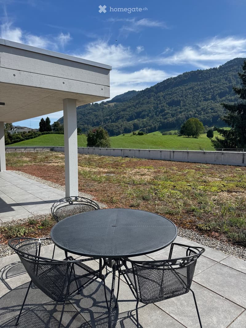 Balcony with a round metal table and two matching chairs, overlooking a green, hilly landscape with trees and a clear blue sky.