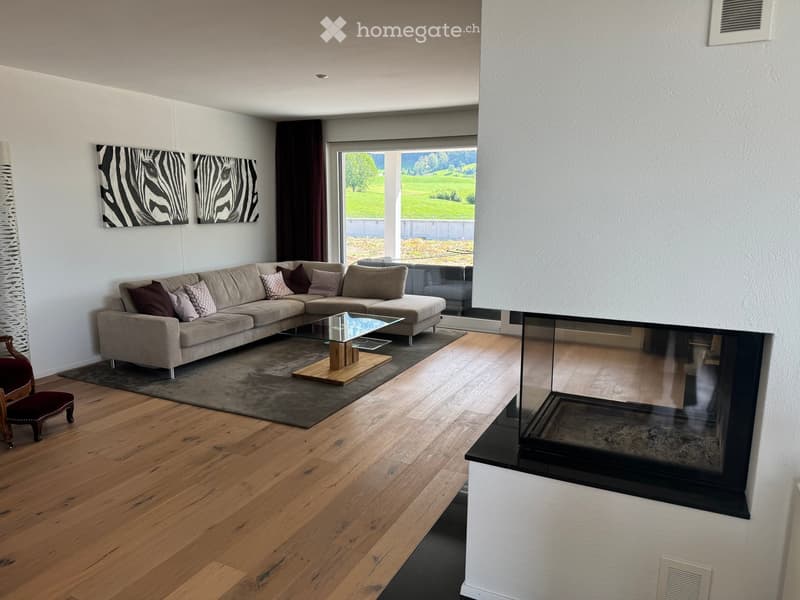 Modern living room with light wooden flooring, a sectional sofa, a glass coffee table, a fireplace, and large windows offering a scenic view.