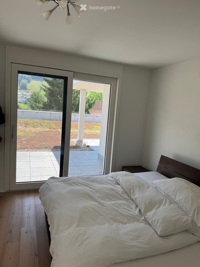 Bright bedroom with wooden flooring, large sliding glass doors leading to a patio, a neatly made bed with white bedding, and a nightstand.