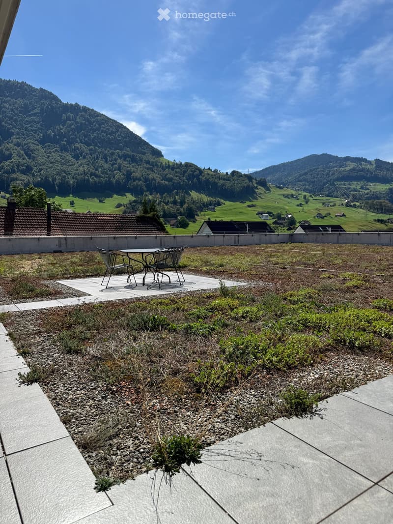 A rooftop terrace with a small seating area, featuring a table and chairs, surrounded by a low green roof and a scenic mountain view.
