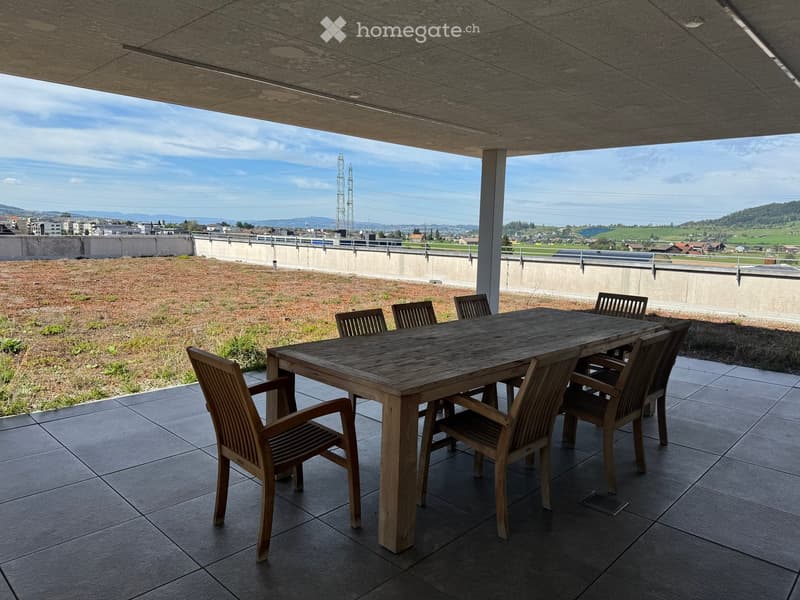 Wooden table and chairs on a tiled balcony with a view of a cityscape and green fields.