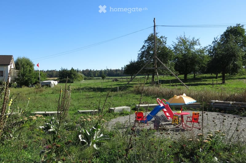 Outdoor play area with a slide, colorful chairs, a small table, and an umbrella, surrounded by grass and wildflowers, with a wooden fence and a house in the background.
