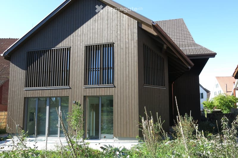 Modern two-story house with dark wooden siding, large windows, and a sloped roof. Features include a covered entrance and surrounding greenery.