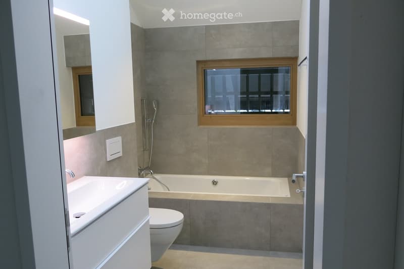 Bathroom. Modern bathroom with a bathtub, toilet, sink, and shower head. Grey tiled walls, a window above the bathtub, and white cabinetry.