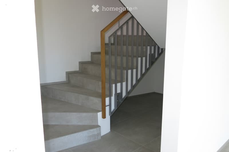 Modern staircase with concrete steps, wooden handrail, and metal balustrade, leading upwards in a well-lit hallway.