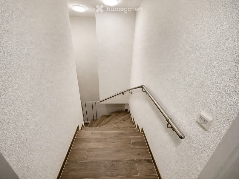 Modern staircase with wooden steps and metal handrails, white textured walls, recessed ceiling lights, and a light switch on the wall.