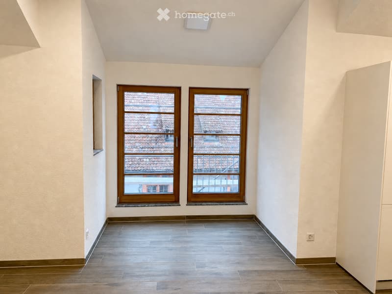 Empty room with light-colored walls, wooden floor, and two windows with wooden frames showing a view of a tiled roof.