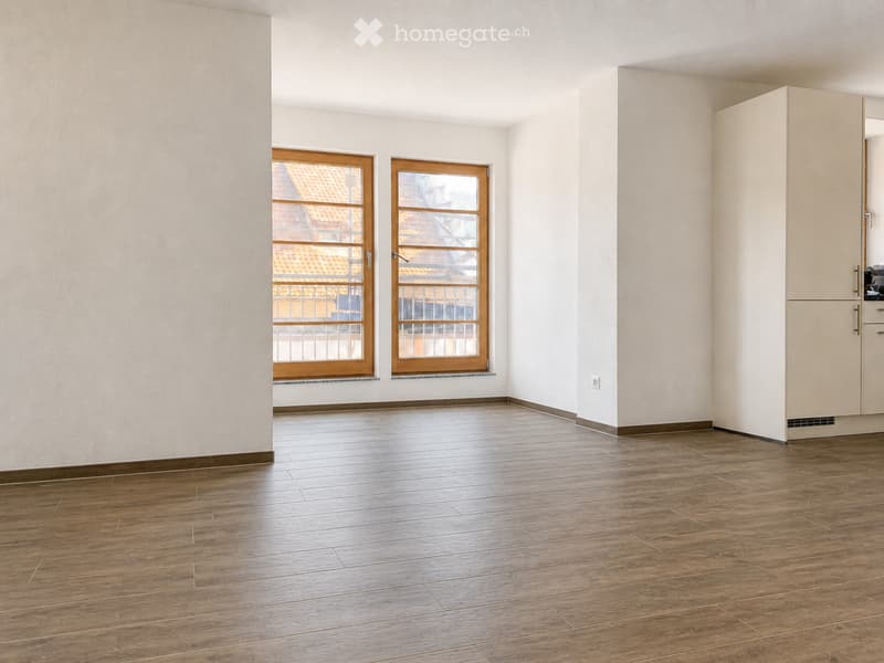 Empty room with wooden flooring, white walls, and two windows with wooden frames. A small cabinet is visible on the right side.