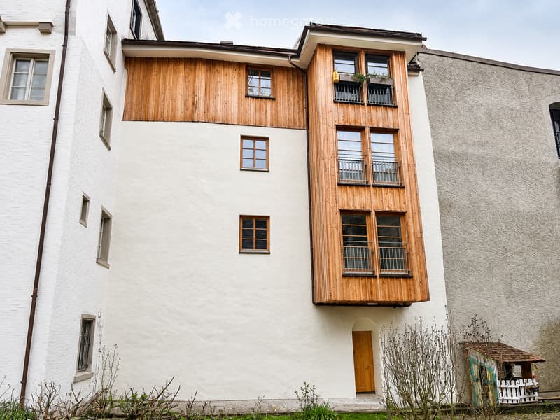 A multi-story residential building with white walls, wooden balconies, and multiple windows. The building features a mix of modern and traditional architectural elements, including a wooden entrance door and small garden area.