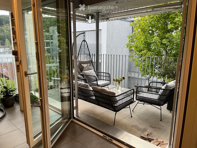 Balcony with a sofa, chairs, a small table, a vase with flowers, and potted plants. The balcony has a wooden floor and is enclosed with a metal railing.