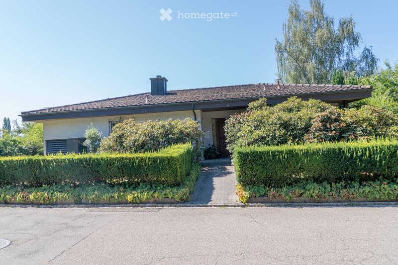 Two-story house with a tiled roof, beige exterior, and a small chimney. The house is surrounded by well-maintained hedges and trees, with a paved pathway leading to the entrance.