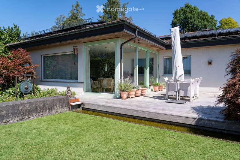 A modern house with a spacious wooden deck, outdoor dining set, potted plants, and a well-maintained lawn.