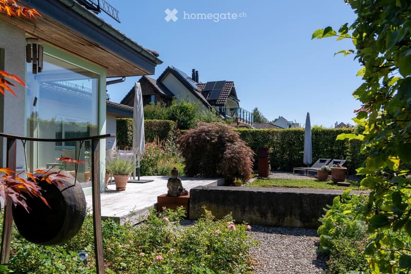 Well-maintained garden with a variety of plants, a stone bench, a Buddha statue, and a modern house with large windows and a wooden deck.