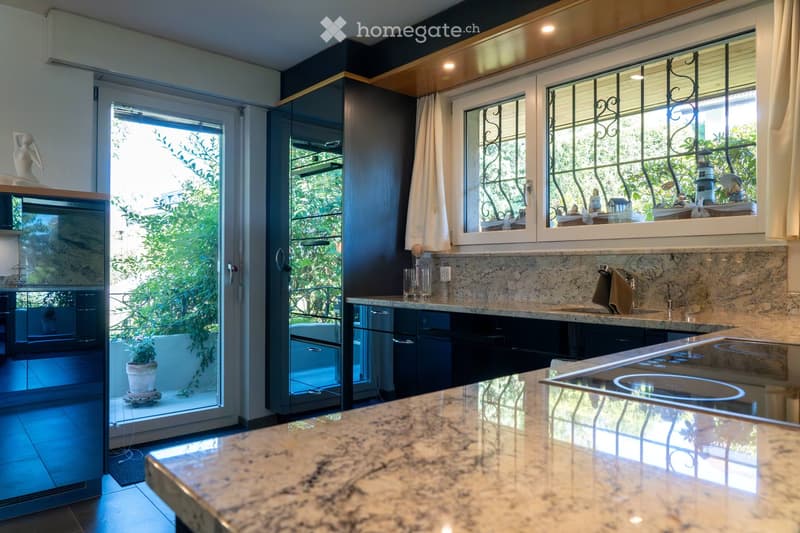 Modern kitchen with granite countertops, stainless steel appliances, large windows with decorative iron bars, and a sliding glass door leading to a balcony.