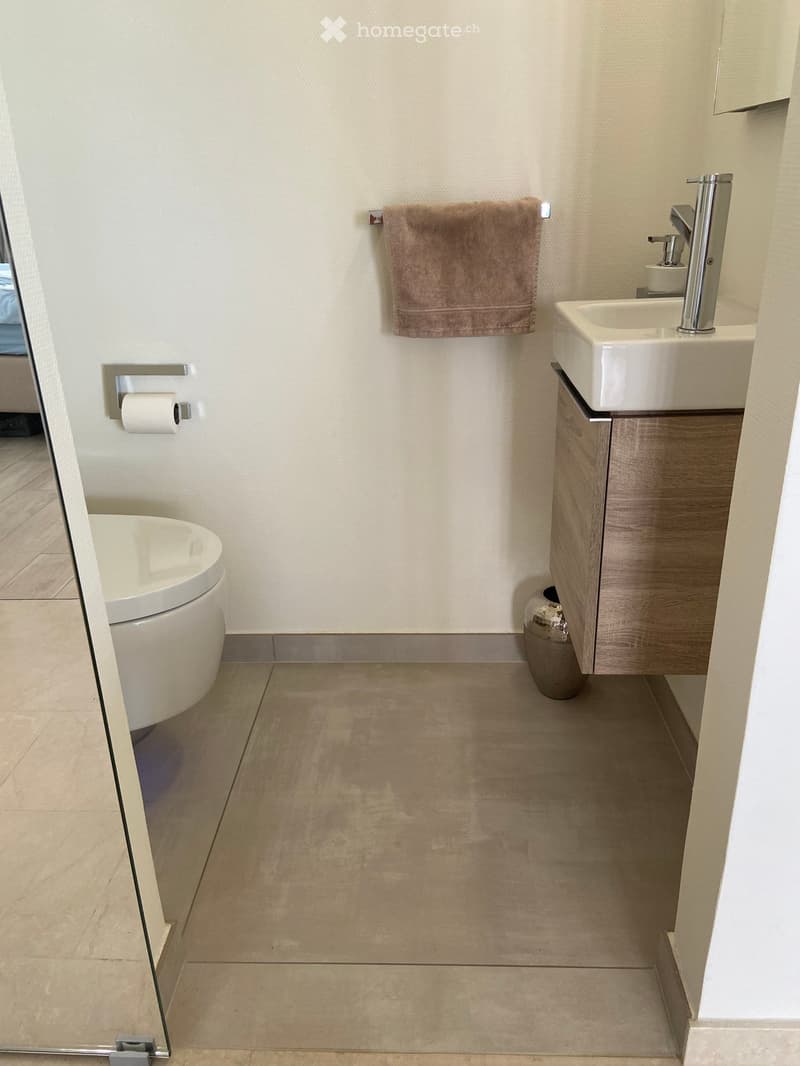 Modern bathroom with a toilet, sink, and wooden cabinet. Beige tiled floor and walls, with a towel hanging on the wall and a small decorative vase on the floor.