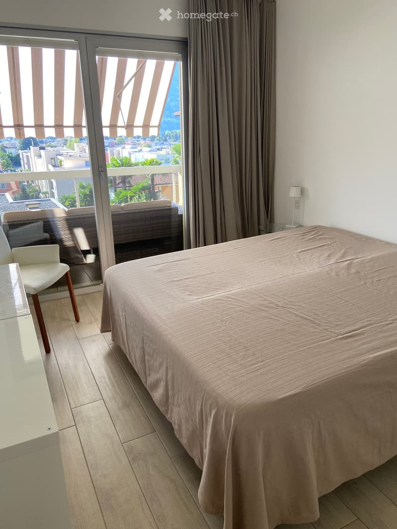 Modern bedroom with a large bed, a white chair, a small table with a lamp, and large windows with curtains offering a scenic view.