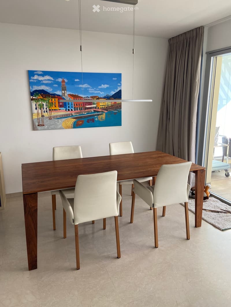 Modern dining room with a wooden dining table, four white chairs, and a large painting on the wall. The room features large windows with curtains and a view of the outside area.