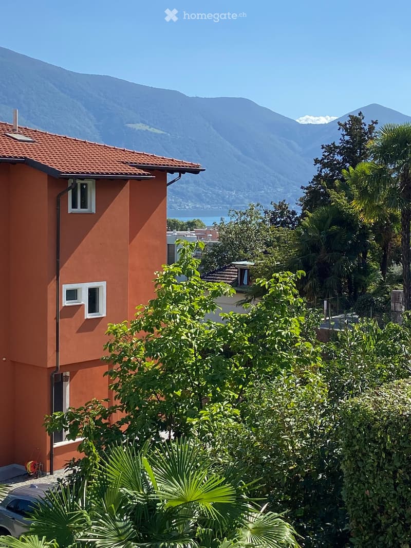 A two-story orange house with white-framed windows, surrounded by lush greenery and palm trees, overlooking a scenic lake and mountain range.