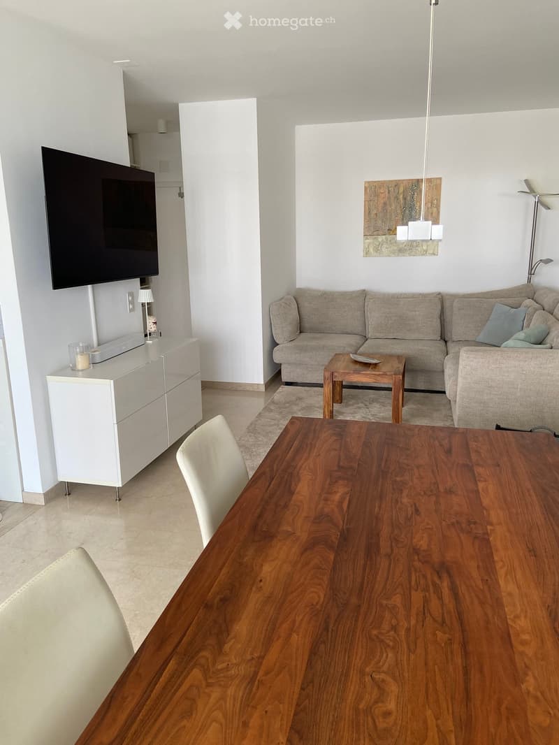 Modern living room with a grey sofa, wooden coffee table, dining table, white cabinet, and a flat-screen TV mounted on the wall.