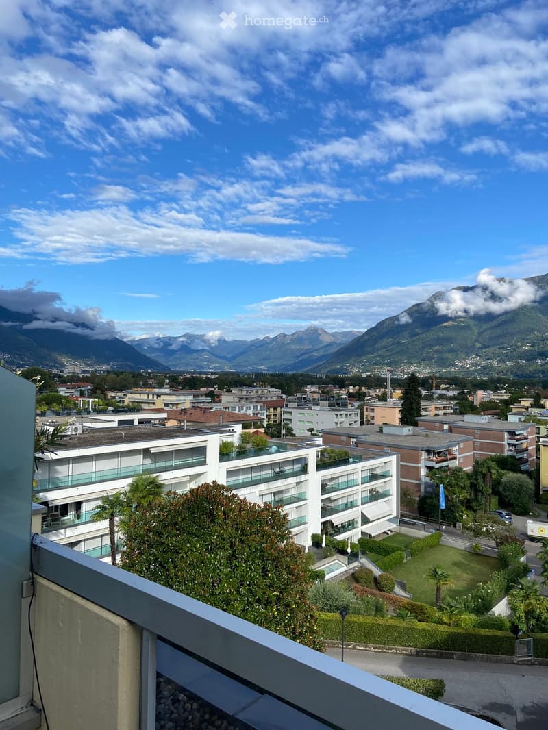 Balcony overlooking a cityscape with modern apartment buildings, greenery, and distant mountains under a partly cloudy sky.