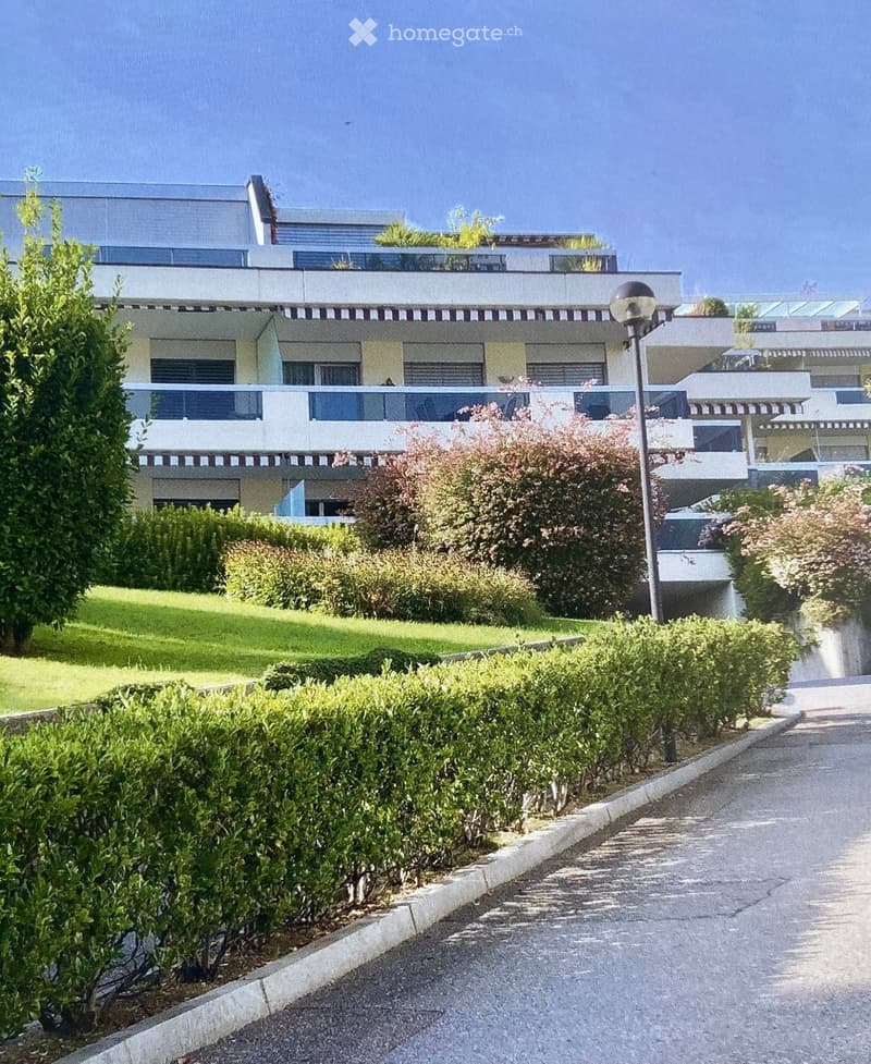 Modern multi-story residential building with balconies, surrounded by well-maintained greenery and a paved pathway.