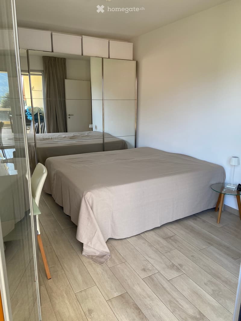 Modern bedroom with a large bed, a wardrobe with mirrored doors, a chair, a small table with a lamp, and a glass door leading to a balcony.