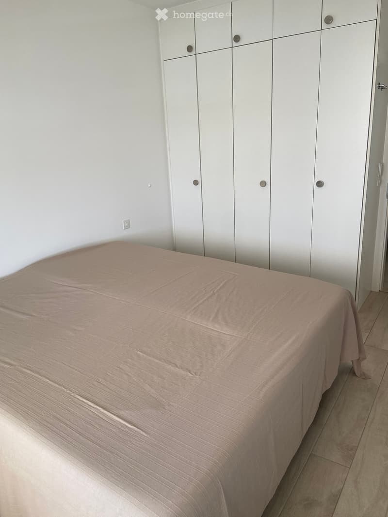 A modern bedroom with a neatly made bed covered in a light pink duvet, positioned against a white wall. A large white wardrobe with multiple doors is visible to the right of the bed.