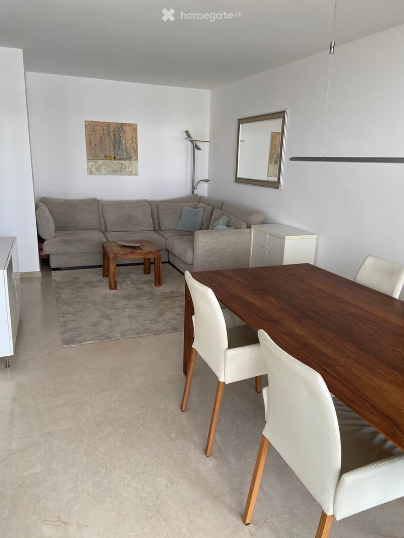 Modern living and dining area with a grey sectional sofa, wooden coffee table, dining table with white chairs, a mirror, and a floor lamp.