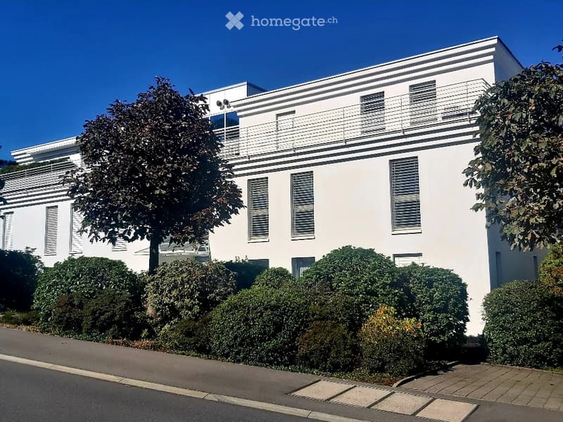 Modern two-story white building with large windows, balconies, and trimmed hedges in front.