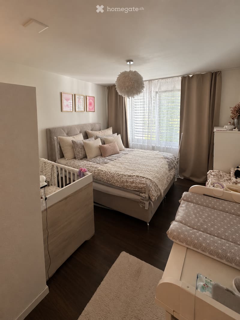 Cozy bedroom with a large bed, multiple pillows, a blanket, a baby crib, a baby chair, a soft rug, and a decorative lamp hanging from the ceiling. The room features large windows with curtains and framed artwork on the wall.