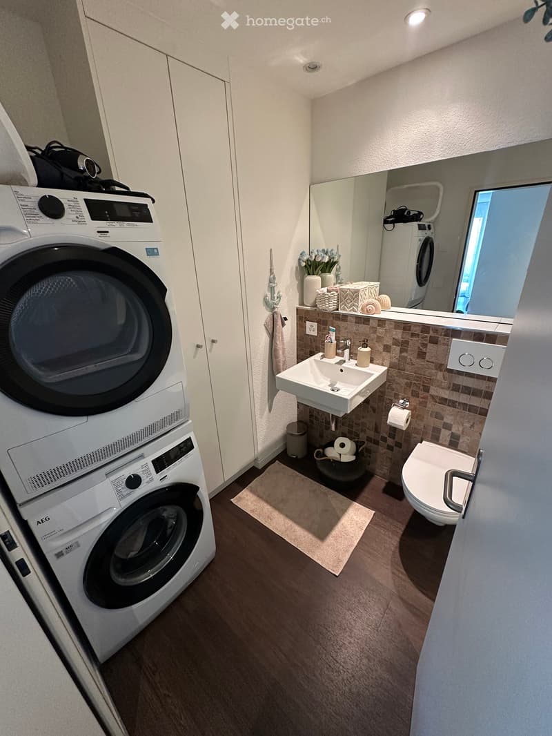 Modern bathroom with a sink, toilet, washing machine, dryer, and a mirror. The room features a tiled backsplash, a small window, and a wooden floor.