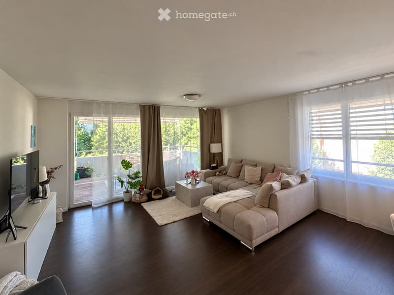 Modern living room with hardwood flooring, large windows, and a balcony. Features a beige sectional sofa, coffee table, TV, and decorative plants.