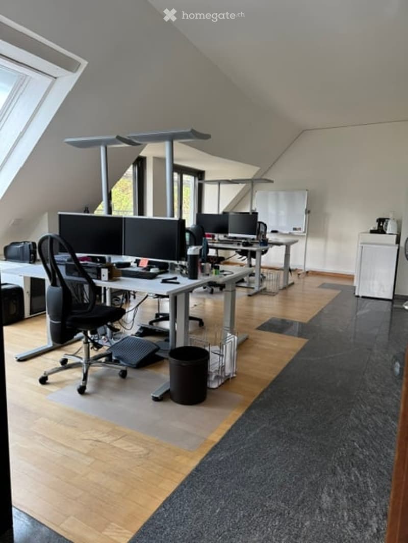 Modern office space with multiple desks, ergonomic chairs, computer monitors, keyboards, and a whiteboard. The room features a mix of wooden flooring and carpeted areas, with large windows providing natural light.