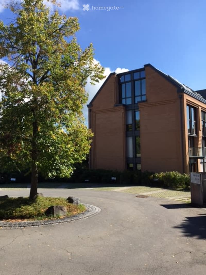 Modern multi-story building with red brick facade and large windows, surrounded by a paved area and greenery.