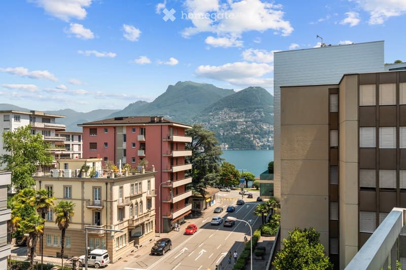 A scenic view of a lakeside town with multi-story buildings, a road with cars, and a backdrop of mountains and a lake.
