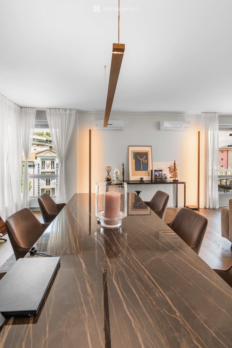 Modern dining area with a long marble table, six brown leather chairs, a laptop, a candle in a glass holder, a vase, picture frames, books, and a sculpture on a side table.