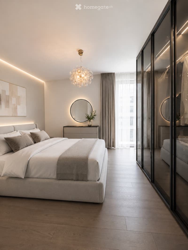 Modern bedroom with a large bed, nightstand, round mirror, potted plant, and a chandelier. The room features wooden flooring and sleek, contemporary decor.