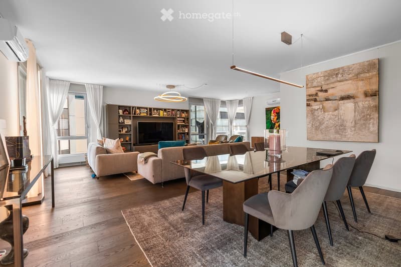 Modern living room with a large glass dining table, grey chairs, beige sofas, a bookshelf, and a television. The space features large windows with white curtains, wooden flooring, and contemporary decor including artwork and lamps.